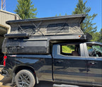 A black pickup truck parked outdoors in front of a building, with a Cascadia Overland Apex mounted in thet ruck bed. Trees and blue sky are visible in the background.